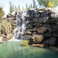 A natural-looking rock waterfall with multiple cascades flowing into a greenish pool, surrounded by trees and grass.