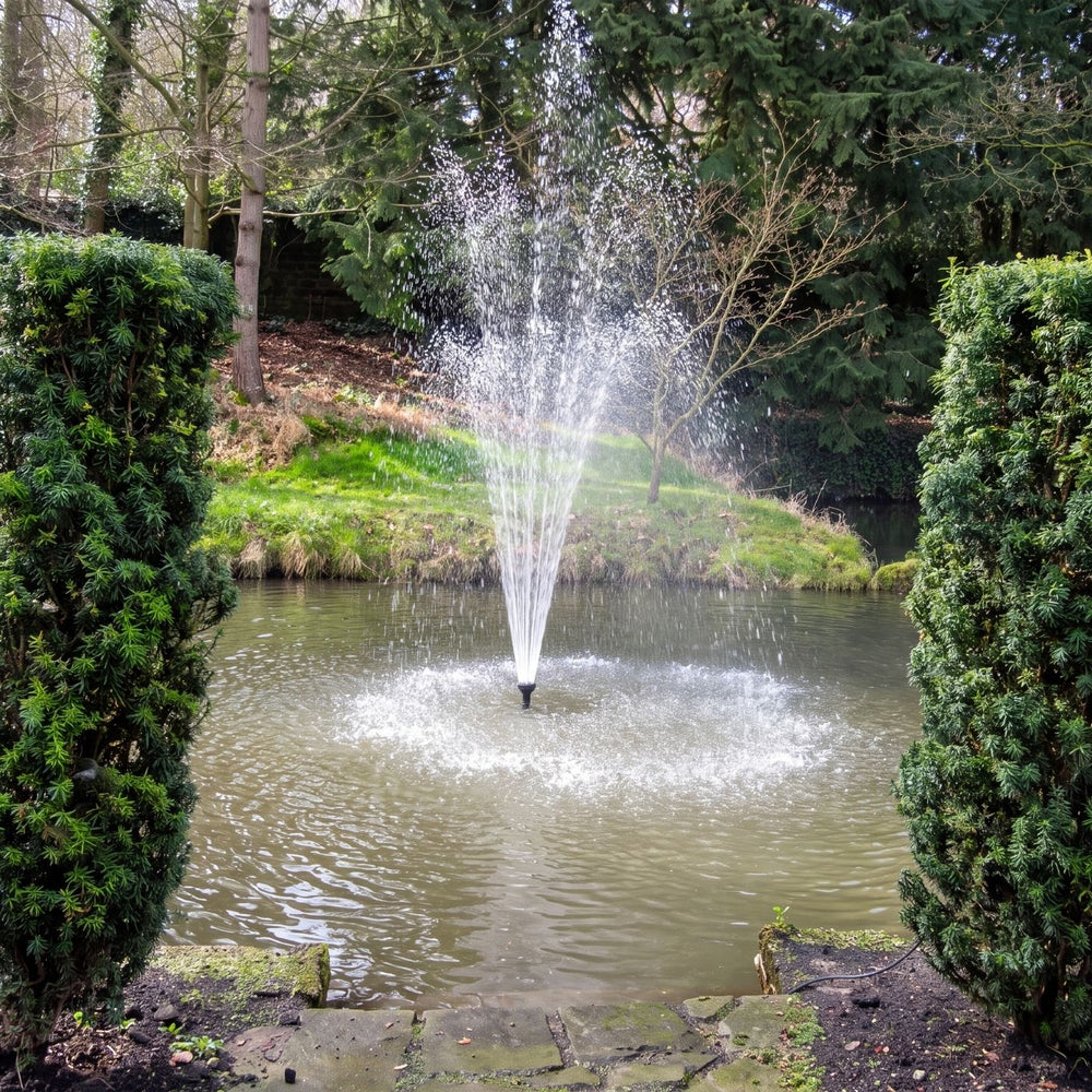 A small garden pond fountain spraying water upward, centered in a landscaped yard with trimmed green hedges, trees, grass, and a stone path in the foreground.