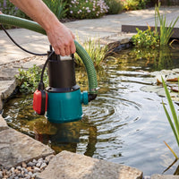 A submersible pond pump with a green hose connected to a garden pond, being operated by a person's hand. The pump features a teal body, a black top section, and a red control box.