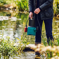 A person standing in shallow water near a pond or stream holds a teal and black submersible pump with a red float. The background shows green plants and reflective water in an outdoor garden setting.