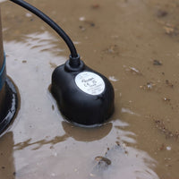 A black submersible pump with an automatic floor sensor featuring EVAK sitting in muddy water.