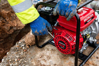 A red Honda WB20 water pump being started by a worker wearing blue work gloves and a high-visibility yellow jacket on a dirt construction site.
