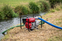 A Honda portable water pump with a red engine and black metal frame, positioned outdoors next to a stream with a green hose connected to it and a blue hose extending to the right.