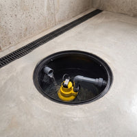 A yellow and gray submersible pump installed inside a circular drain opening in a concrete floor, with a black floor drain and textured beige walls in the background.