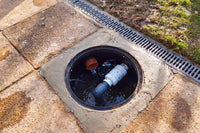 A circular concrete access opening showing a black drain or pipe assembly inside, set into a stone paved surface with a metal grate and grass visible at the top right.