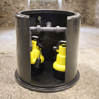 A submersible sump pump system inside a cylindrical black metal casing, with two bright yellow pumps visible through an open cutout, set on a concrete floor against a stone wall background.