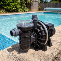 A black and gray pool pump with a clear filter lid sits on a concrete pool deck next to a blue swimming pool.