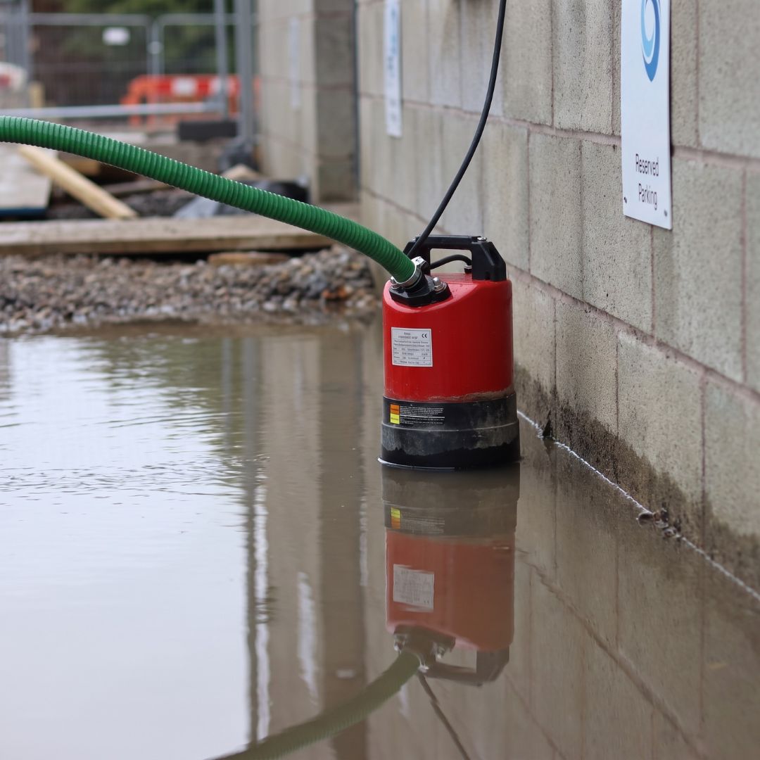 SPK530 submersible puddle pump connected to a green hose, installed in shallow water along a concrete block wall.
