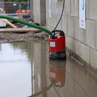 SPK530 submersible puddle pump connected to a green hose, installed in shallow water along a concrete block wall.