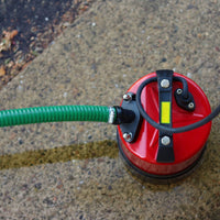 A red puddle pump with a green hose connected, sitting on a wet concrete ground surface.