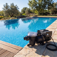 A black swimming pool pump with a clear filter lid sits on a stone patio beside a bright blue swimming pool, with trees and landscaping in the background.