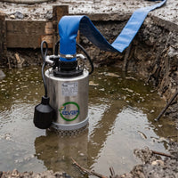 An automatic submersible Evak Drainox site drainage pump with a stainless steel body, blue hose, and black power cord, shown partially submerged in muddy water at a construction site.