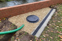 Outdoor drainage system showing a round black manhole cover, a long black metal gutter channel, a green flexible pipe, and light gravel beside a brick retaining wall and water.