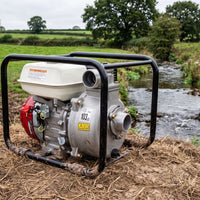 A portable gasoline water pump with a black tubular frame, a silver metal pump body, a white plastic housing, and a red engine cover, positioned outdoors next to a flowing stream with green fields in the background.