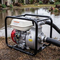 A portable outdoor water pump with a black metal frame, silver pump body, white fuel tank, and red engine housing, positioned on muddy ground beside a flooded pond with water flowing out via a black hose.