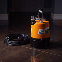 A puddle pump with a bright orange body and black base, shown on a dark tiled floor next to its coiled black power cord. The pump has a visible Tsurumi brand label.