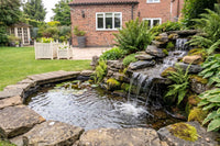 An outdoor backyard pond feature with a small waterfall by APP TPS Pump cascading over stacked natural-looking rocks into a stone-lined pool.