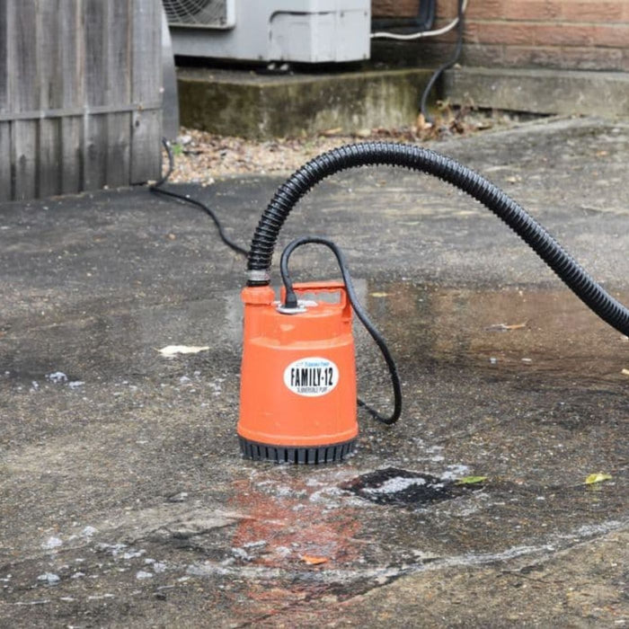 tsurumi family 12 submersible puddle pump with black hose on a concrete surface, with a wooden fence and air conditioning unit in the background.